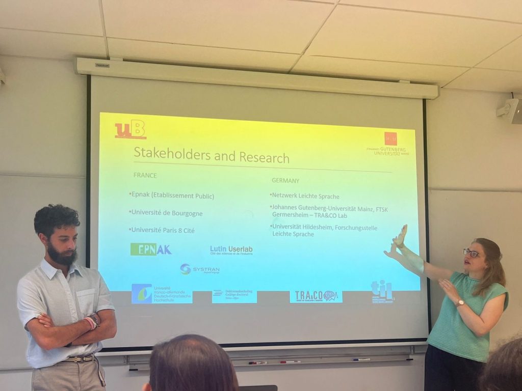 Two presenters standing in front of a slide with information on French and German organizations involved in Easy Language. To the left Léo Anebi in a white T-Shirt and to the left Julia Degenhardt in a turquoise shirt.